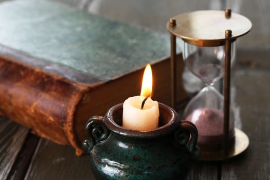 Flighting candle near hourglass and book on old wooden surface