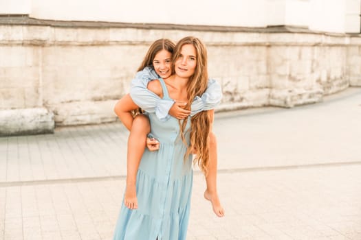 Mother of the daughter walks playing. Mother holds the girl on her back, holding her legs, and her daughter hugs her by the shoulders. Dressed in blue dresses