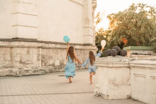 Daughter mother run holding hands. In blue dresses with flowing long hair, they hold balloons in their hands against the backdrop of a sunset and a white building