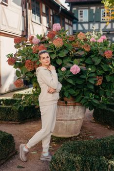 Attractive curly blonde woman walk on the city park street. Girl wear purple hoodie look happy and smiles. Woman make here me gesture standing near pink blooming bush flowers. Happy laughing girl