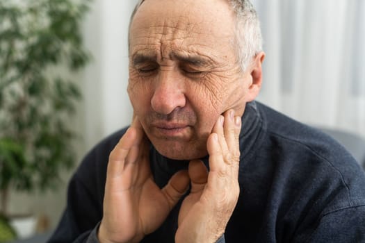 Old man with toothache. Elderly senior man has toothache. Unhappy man face in tooth pain sitting on sofa at home, feel sick unwell. Sad aged man hand holding his chin. Adult suffering toothache.