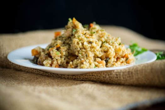 cooked bulgur with vegetables, carrots and dried mushrooms in a plate on a wooden table.