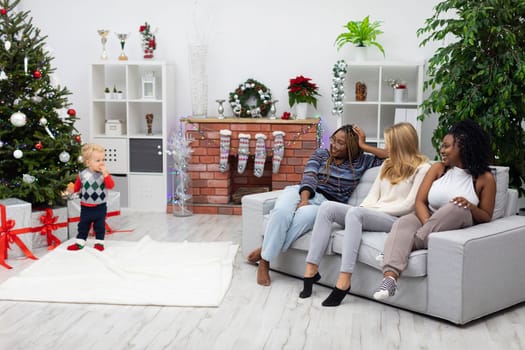 The girls sit on the sofa and look at the two-year-old who is standing by the Christmas tree. Two African women and one European woman sit together side by side. International Christmas.