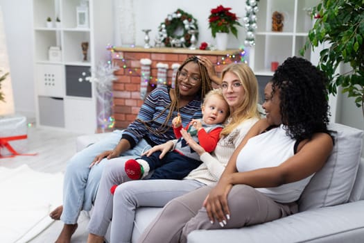 Three friends are sitting on the sofa, two of them are African and one is European. The sister is sitting on the sofa and holding her little brother on her lap.