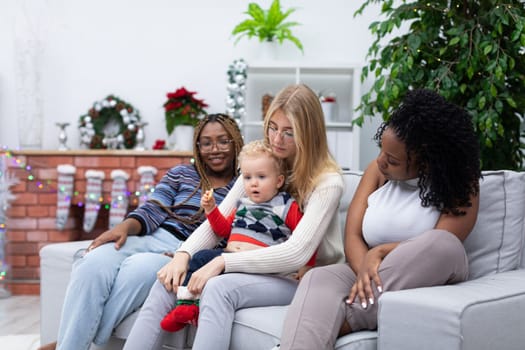 A little boy sits at his sister's lap. Christmas decor of the room. Two dark-skinned friends sit on the sofa.