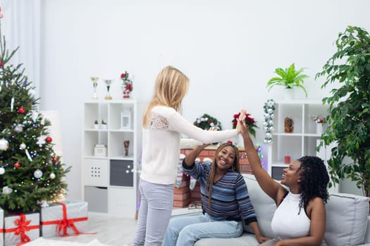 Happy friends greet each other. They high-five each other in a gesture of welcome. The room decorated in the atmosphere of Christmas. African women and a European woman.
