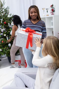A smiling African woman gives a gift package to her European friend. A living room decorated for Christmas.