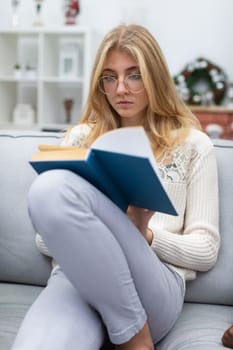 A blonde woman with glasses sits on a sofa and reads a reading. Christmas decoration. A student is studying from a book. A diligent student.