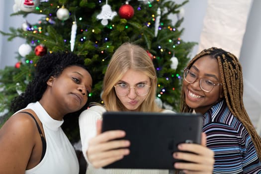 Christmas selfie of friends against the background of a Christmas tree. Funny photos taken with a tablet in a decorated living room at home.