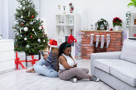 Two friends from Africa sit in the living room and look at their smartphones. Christmas decoration of the living room.