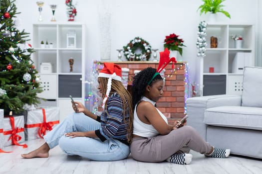 Two friends from Africa sit in the living room and look at their smartphones. Christmas decoration of the living room.