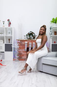 A dark-skinned girl sits on a sofa in a white dress and laughs. She is waiting for invited guests on Christmas Day.