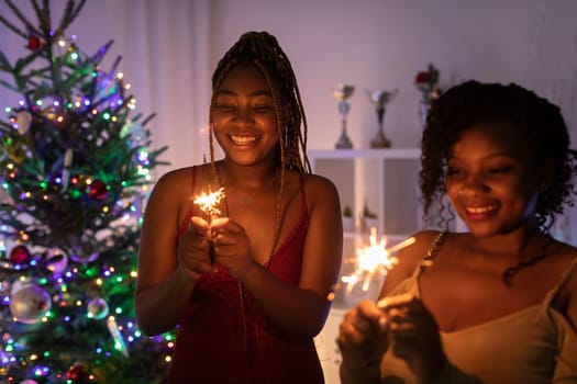 In the background is a Christmas tree with twinkling lights. Two dark-skinned girls hold lit cold fires in their hands.