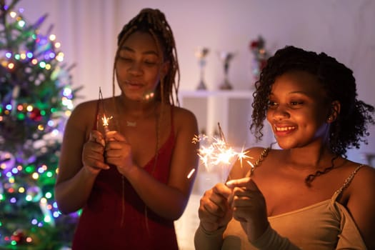 In the background is a Christmas tree with twinkling lights. Two dark-skinned girls hold lit cold fires in their hands.