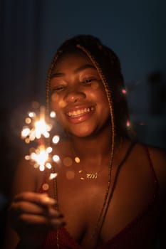 Against a dark background, a dark-skinned girl stands and smiles while holding burning cold fires in her hand. Happy New Year celebration.