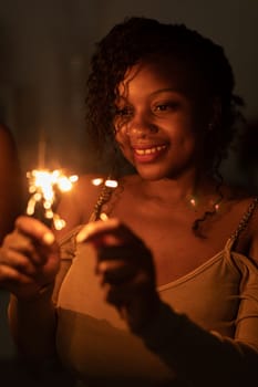 Against a dark background, a dark-skinned girl stands and smiles while holding burning cold fires in her hand. Happy New Year celebration.