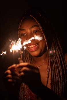 A girl with African beauty during the night of New Year's Eve lit cold fires and holds them in her hand. A happy and smiling student.