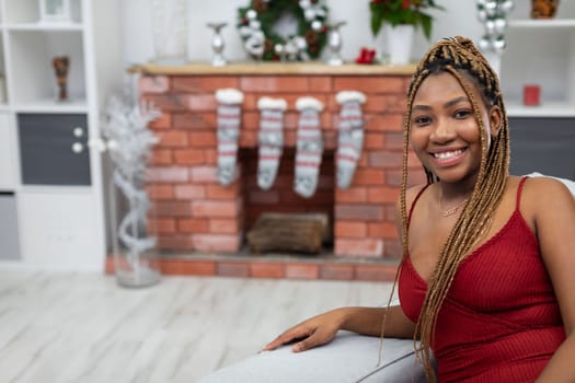 A dark-skinned girl dressed elegantly in a red dress sits smiling on a sofa. The living room decorated for Christmas Eve.