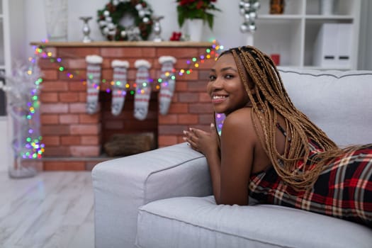 An African woman with braided pigtails lies on a gray sofa. The living room decorated with Christmas decorations. Fireplace with Christmas socks.