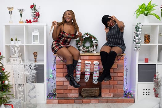 The mischievous dark-skinned sisters sat on the fireplace decorated for Christmas. Christmas socks hang by the fireplace.