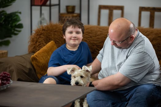 In the living room, a father and son sit on the sofa and stroke their multi-racial dog.