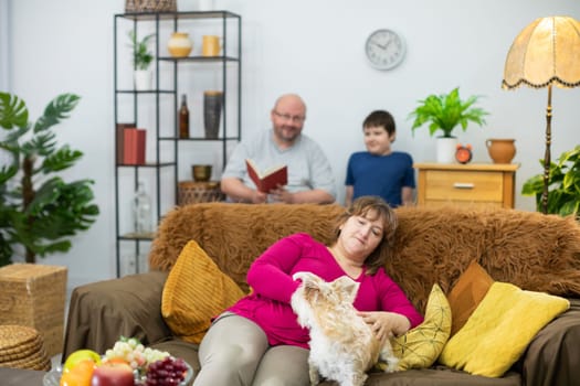 In the living room, the father is reading a book to his son and the mother is playing with the shaggy dog on the sofa.