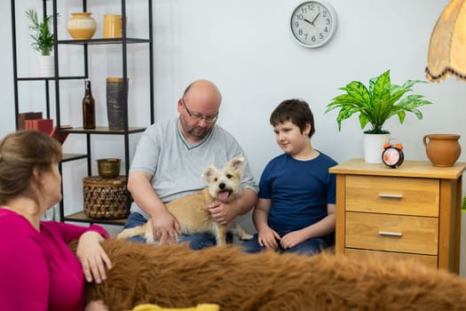 Mom watches from the sofa as father and son play with a shaggy dog in chairs against the wall next to a bookcase and dresser.