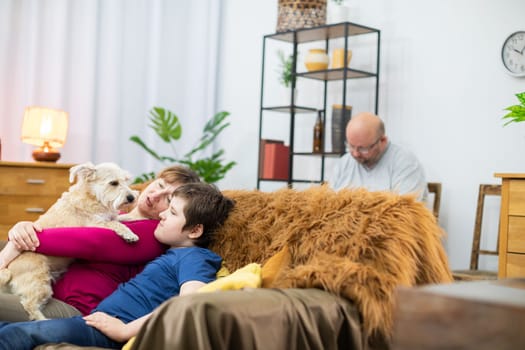 The mother holds the shaggy dog in her arms and the son cuddles with her. The father is quietly reading a book.