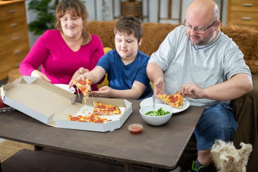 A shaggy dog watches when he too gets his slice. Mother, Father and son help themselves to hot and fragrant slices of pizza from a box.