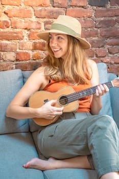 Cheerful young woman playing ukulele on the sofa in a cozy home in boho style. High quality photo