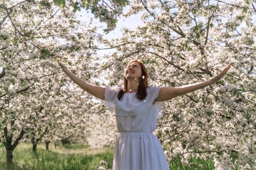 Fantasy woman in long white elegant fashion long dress walks in green spring blossom cherry garden. Happy cheerful girl princess bride. Skirt fabric flies flowing waving in wind motion