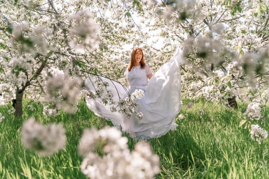 Fantasy woman in long white elegant fashion long dress walks in green spring blossom cherry garden. Happy cheerful girl princess bride. Skirt fabric flies flowing waving in wind motion