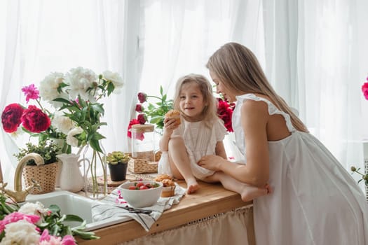 A little blonde girl with her mom on a kitchen countertop decorated with peonies. The concept of the relationship between mother and daughter. Spring atmosphere.