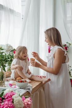 A little blonde girl with her mom on a kitchen countertop decorated with peonies. The concept of the relationship between mother and daughter. Spring atmosphere.