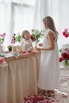 A little blonde girl with her mom on a kitchen countertop decorated with peonies. The concept of the relationship between mother and daughter. Spring atmosphere.