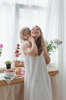 A little blonde girl with her mom on a kitchen countertop decorated with peonies. The concept of the relationship between mother and daughter. Spring atmosphere.