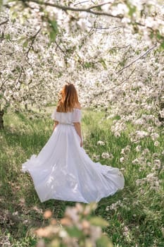 Fantasy woman in long white elegant fashion long dress walks in green spring blossom cherry garden. Happy cheerful girl princess bride. Skirt fabric flies flowing waving in wind motion