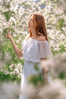 Woman cherry orchard. A happy woman in a long white long dress walks through the green spring blooming cherry garden. Happy cheerful princess bride. The fabric of the skirt flutters in the wind