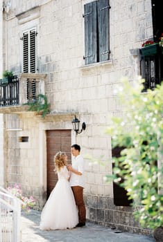 Groom holds the bride hands standing near the old stone house with shutters on the windows. High quality photo