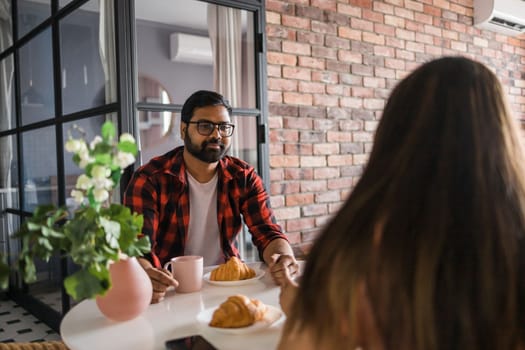 Young diverse loving couple eating croissant and talks together at home in breakfast time. Communication and relationship