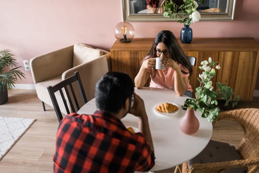 Young diverse loving couple eating croissant and talks together at home in breakfast time. Communication and relationship