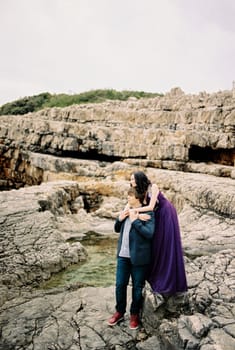 Woman in a lilac dress hugs man from behind standing on the rocks. High quality photo