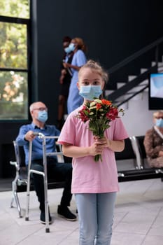 Little girl standing in hospital looby area while holding bouqet of flowers during checkup visit examination. Diverse people wearing protective medical face mask to prevent infection with coronavirus