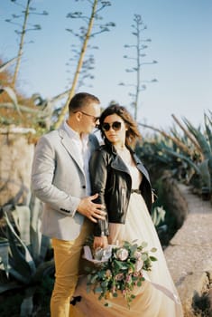 Groom in sunglasses hugs bride in a leather jacket on a stone path. High quality photo