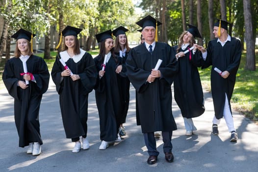 A group of graduates in robes with diplomas in their hands walk outdoors. Elderly student