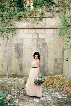 Bride in a dress stands near a stone wall and looks at a bouquet in her hand. High quality photo