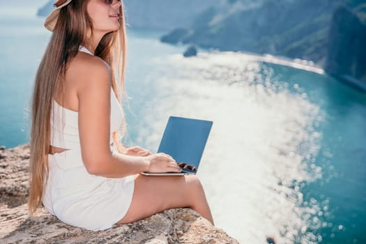 Successful business woman in yellow hat working on laptop by the sea. Pretty lady typing on computer at summer day outdoors. Freelance, travel and holidays concept.