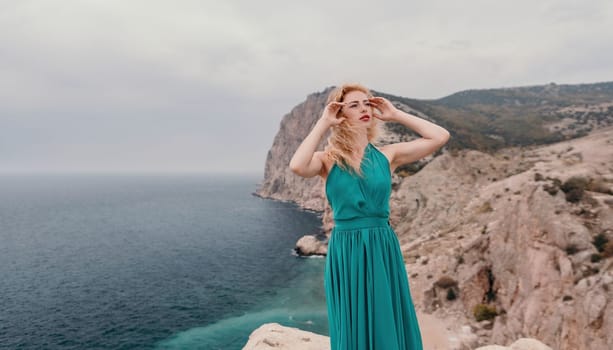 Side view a Young beautiful sensual woman in a mint long dress posing on a volcanic rock high above the sea during sunset. Girl on the nature on overcast sky background. Fashion photo