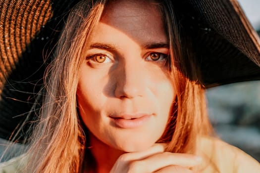 Portrait of happy young woman wearing summer black hat with large brim at beach on sunset. Closeup face of attractive girl with black straw hat. Happy young woman smiling and looking at camera at sea