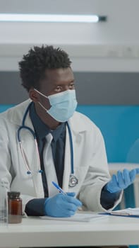 Medic consulting elder patient with face mask and gloves for coronavirus protection at healthcare facility. Young doctor helping senior woman during pandemic at checkup appointment